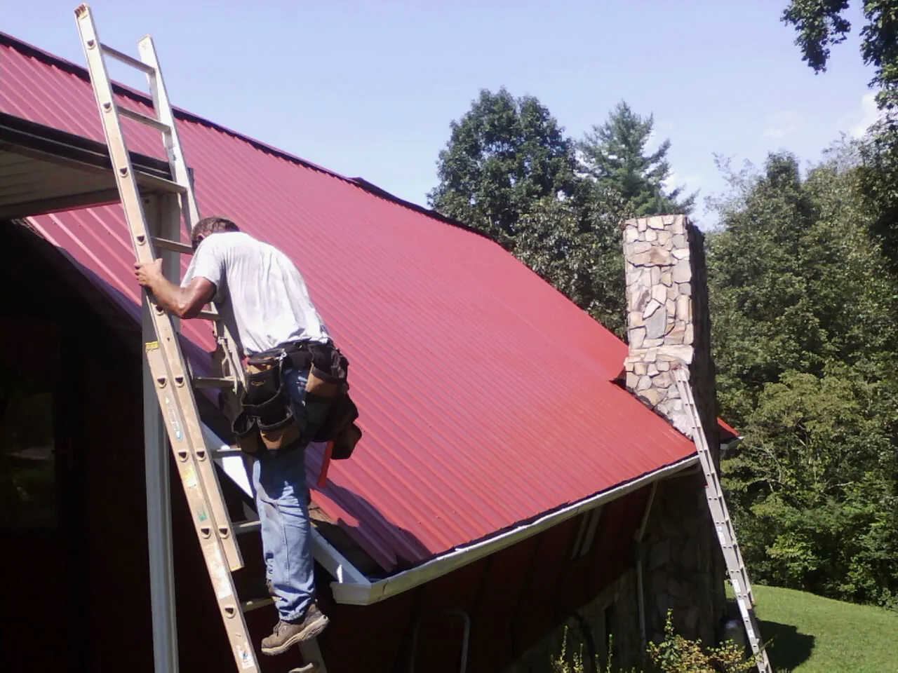 Roofer on a ladder beside a red metal roof and stone chimney — Soffit & Fascia Repair by Roofing Brothers in Butte Falls